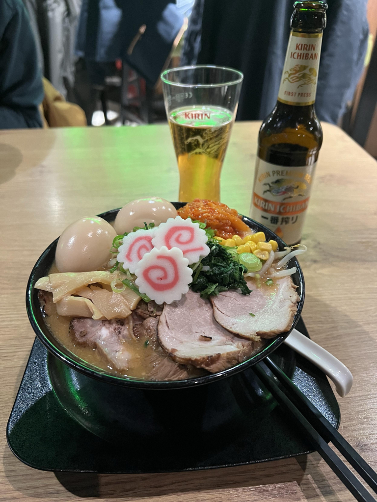 Eine herrliche Schüssel Ramen (japanische Nudelsuppe). Garniert mit 2 Eiern, Mais, Gemüse, Kimchi, Schweinefleisch Scheiben, Bambussprossen, Mais und Gemüse. Dahinter ein Glas Kirin Bier und die zugehörige Flasche