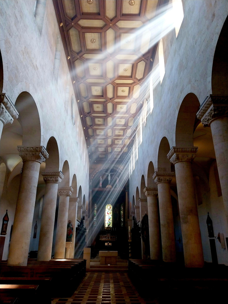 Der Innenraum einer Kirche, oben eine hölzerne Kasettendecke, rechts und links Säulengänge mit Rundbögen, kaum erkennbar Kirchenbänke, die ins Dunkel gehüllt sind, von rechts oben dringen Lichtstrahlen durch kleine rechteckige Fenster quer durch den Raum nach links unten .
The interior of a church, above a wooden coffered ceiling, to the right and left colonnades with round arches, barely visible church pews shrouded in darkness, from the top right rays of light penetrate through small rectangular windows across the room to the bottom left.