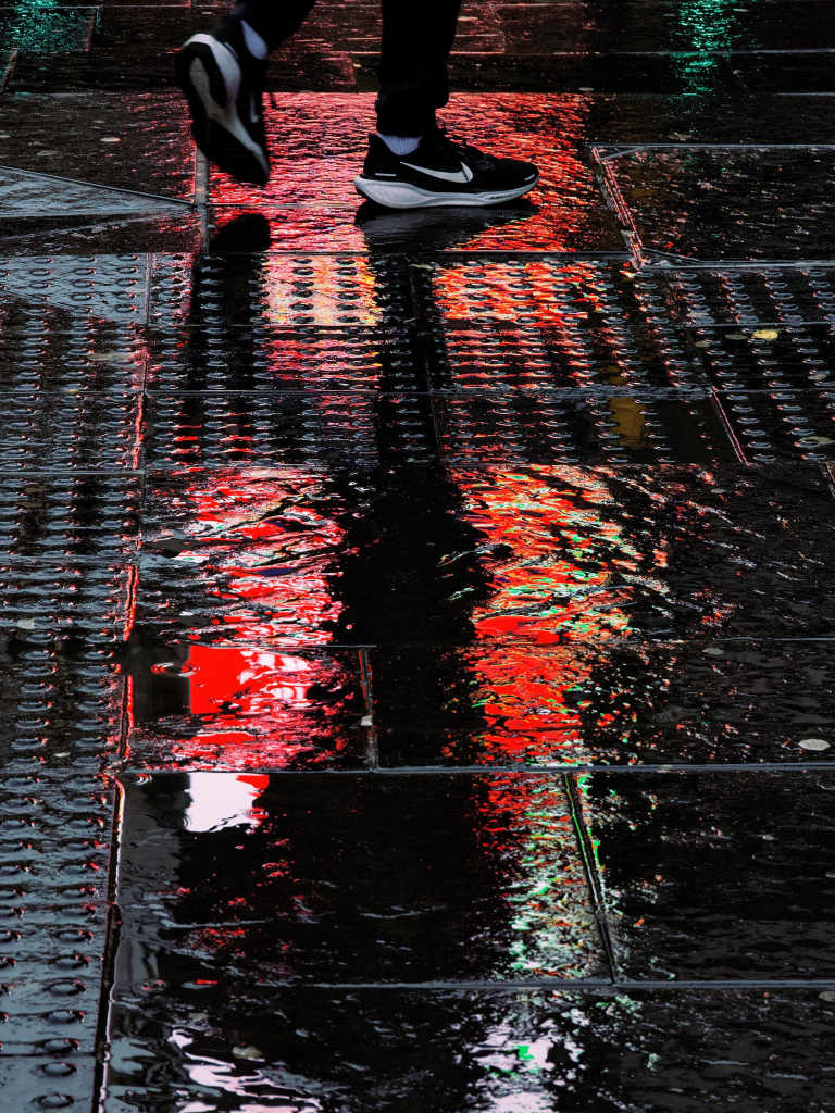 City lights reflecting off the wet street as a pedestrian wearing Nike running shoes crosses the road in Glasgow city centre