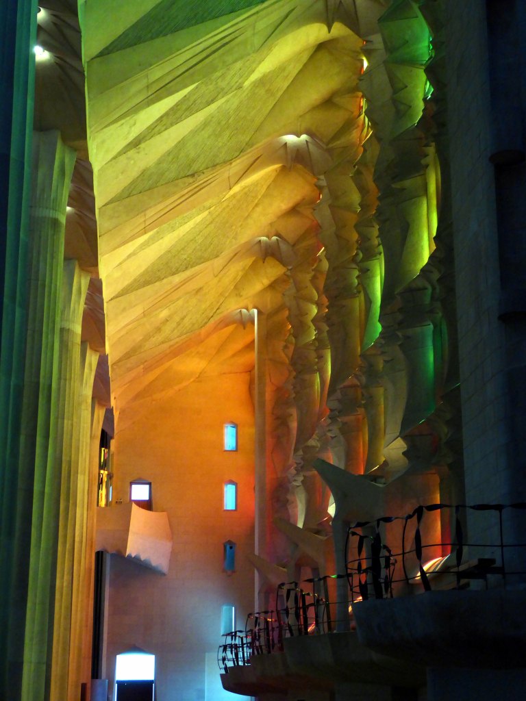 Die Sonne scheint durch die bunten Fenster der Sagrada Família und tauch Decke, Wände und Säulen in bunte Farben. Das gesamte Bild ist von der hinteren Wand bis zum Vordergrund ein Farbverlauf von orange, über gelb bis grün.