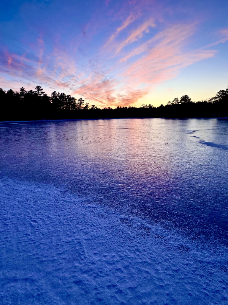 Photo of sunset as viewed from across a frozen lake.  Snowy shoreline in immediate foreground.  Smooth ice on lake reflecting orange, yellow, and red colors of sunset above trees.  Blue sky overhead. 