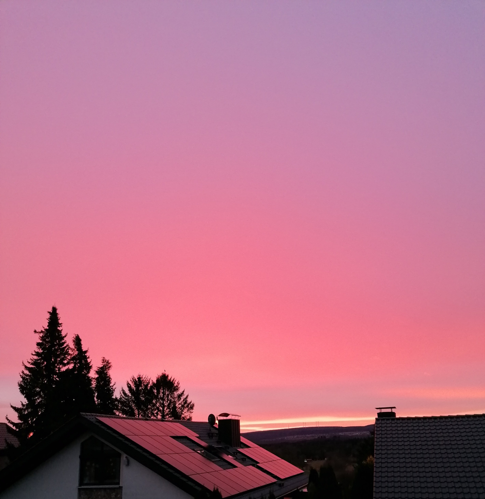 Der Morgenhimmel auf dem Bild zeigt ein beeindruckendes Farbspiel aus intensiven Rosa- und Violetttönen, die sich sanft vermischen. Der Horizont leuchtet in einem helleren Orange, was den Übergang zwischen Nacht und Tag markiert. Die Silhouetten von Häusern und Bäumen im Vordergrund heben sich dunkel gegen den farbenfrohen Himmel ab, wodurch die Szene besonders stimmungsvoll wirkt.

