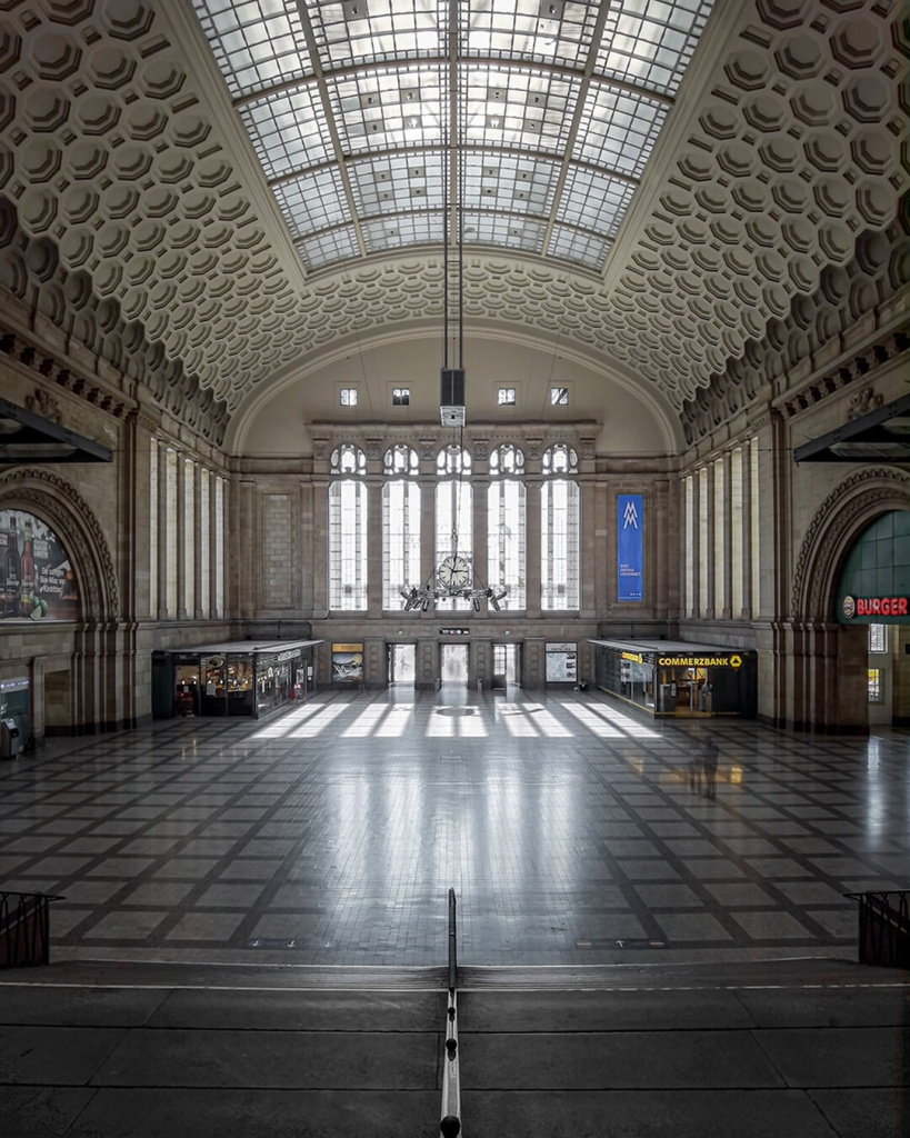 The image shows the central main hall of Leipzig’s central station. The hall is characterized by its impressive architecture: a vaulted ceiling with a geometric pattern of coffers and large glass panels that let in natural light. The floor features a checkerboard pattern of dark and light tiles. In the background, large arched windows allow additional light into the space. On the left and right sides of the hall, shops are visible, including a “Commerzbank” branch and a fast-food restaurant. A chandelier hangs in the middle, emphasizing the historical ambiance.