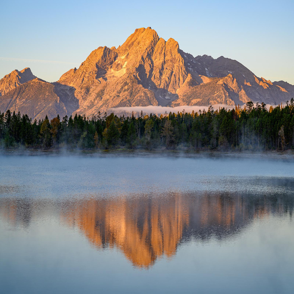 Color photo.  Mount Moran, in Grand Teton National Park, lit by reddish early morning light.  It is reflected in the misty waters of Colter Bay, on the far side of which is a tree lined shore.