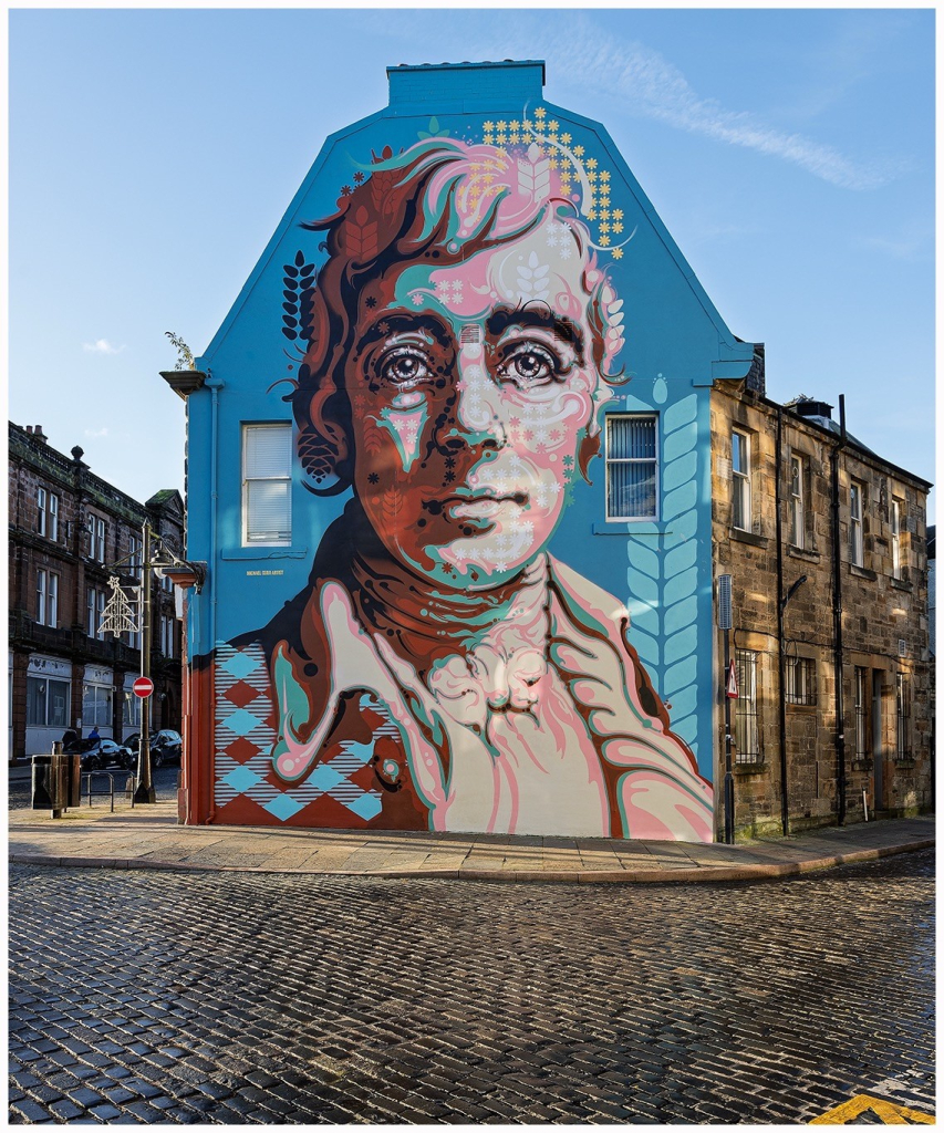 A vibrant mural depicting a stylized portrait of a Robert Burns on the gable end of a building. The mural features bold colors and intricate patterns, set against a blue background. The street is paved with cobblestones, and there are nearby buildings.