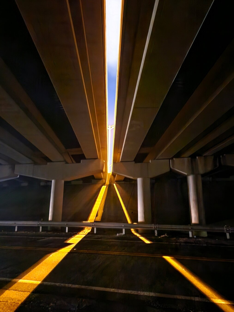 Beneath a highway bridge overpass,  looking up at the gap between the two directional lanes above. Yellow highway lighting illuminates in the night sky and slips through the gap underneath creating an X of yellow in the darkness below.