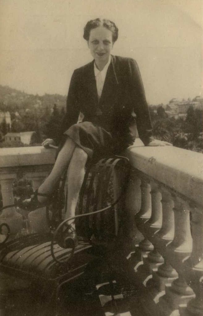 Vintage photograph of a woman sitting on an ornate railing, smiling, dressed in a formal suit.