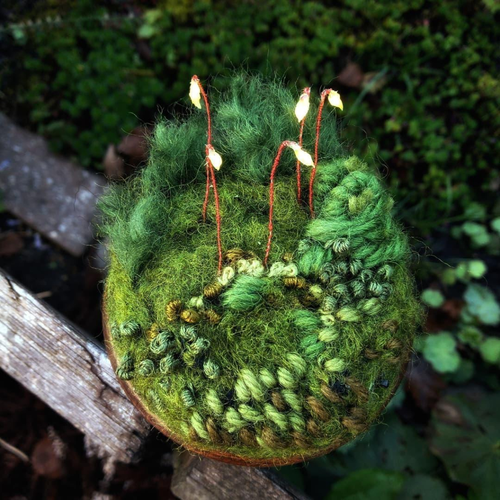 A tiny artwork: lighted silicone sporophytes on an embroidered circular mossy background. The artwork is placed on a piece of wood above a mossy and overgrown forest floor.