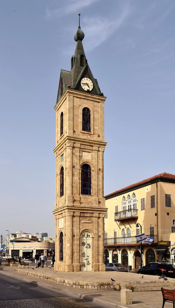 The image depicts a clock tower, specifically the Haifa Clock Tower in Israel. It features a tall, stone structure with a clock face and a distinctive architectural design. The surrounding area includes buildings and streets, suggesting an urban environment. The sky is clear, indicating a bright day.

Image Credits: Wikimedia / Bahnfrend / CC BY-SA 4.0