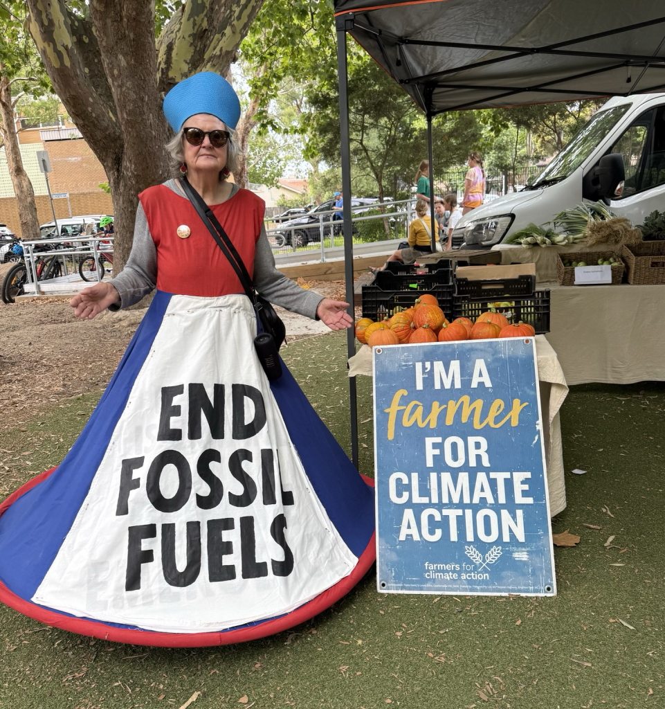 Standing in front of a vegetable stall at a local farmers market: a woman (Sybil Disobedient) wearing a costume with a large blue hoop skirt with a large white panel with the words “END fossil fuels“ standing next to a sign that the stall Owner had, which reads “I’m a farmer for climate action“