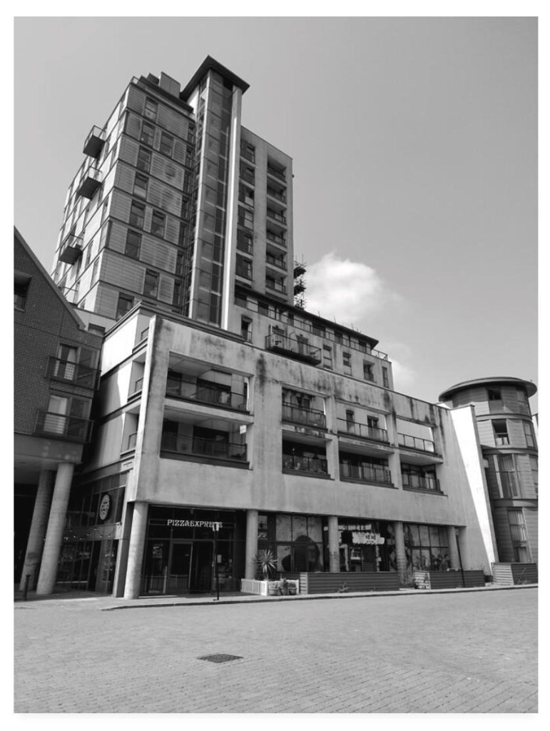 Black and white portrait photograph showing a modern steel and glass block of flats with a pizza restaurant at the base.  In Ipswich, England, if you were wondering.