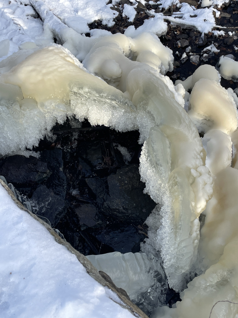 Looking down from the top of the dam into the ice pile formed from the flowing water. While the outer surface of the ice is smooth and slightly yellowish, the inner part where the water splatters is a delicate crystal matrix. It looks like a cut open geode. The wet rocks of the stream bed are seen through this crystal lined opening. 