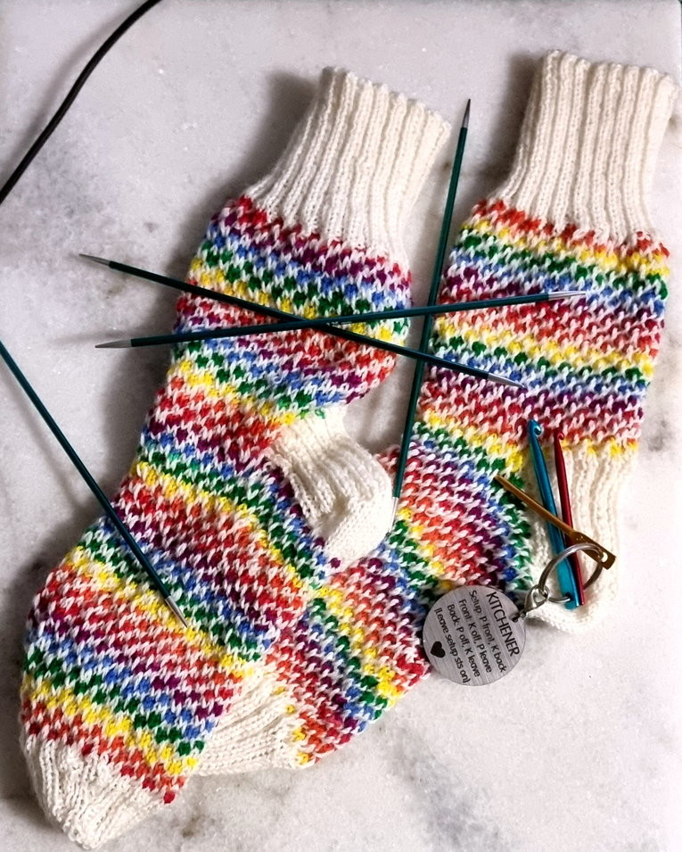 Pair of handknitted socks. Cuff, heel, and toe in cream. Body of sock rainbow stripes with cream between each stitch creating a diamond pattern.
Also in picture is a keyring etched with the instructions for kitchener stitch (used when joining toe).