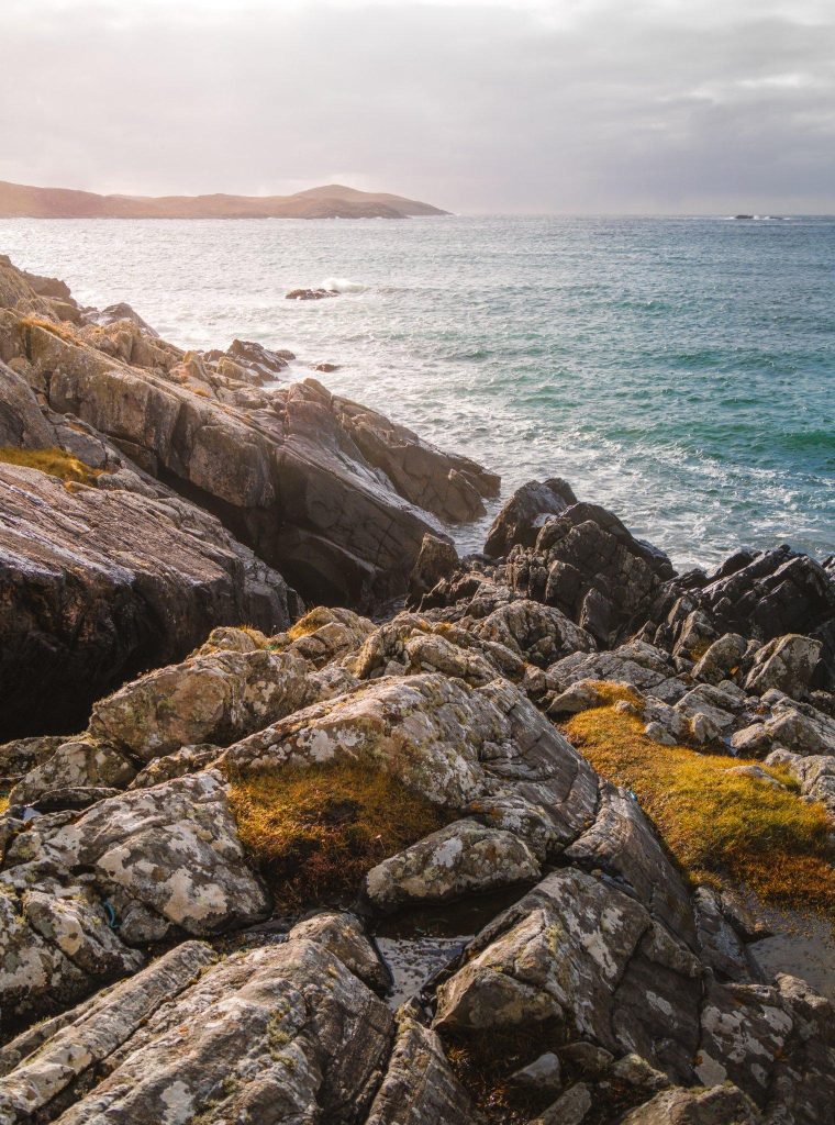 A rocky seascape with azure and blue seas under a cloudy sky, with sun glare coming from the left. Large boulders are in the foreground.