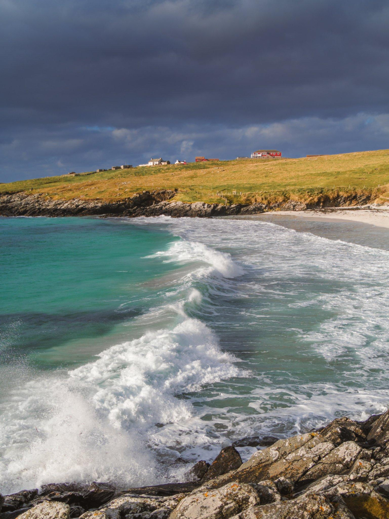 Saturated hues on a beach. White and red cottages are in the background on a hill ending with rocky cliffs, while a white, foaming wave runs from left to right, creating a leading line to the cottages.