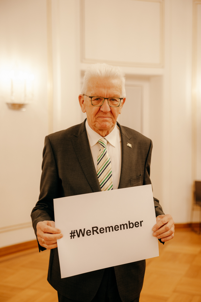 Ministerpräsident Kretschmann mit einem Schild in den Händen zum Gedanken an den 80. Jahrestag der Befreiung des KZs in Ausschwitz. Auf dem Schild steht "#Weremember"