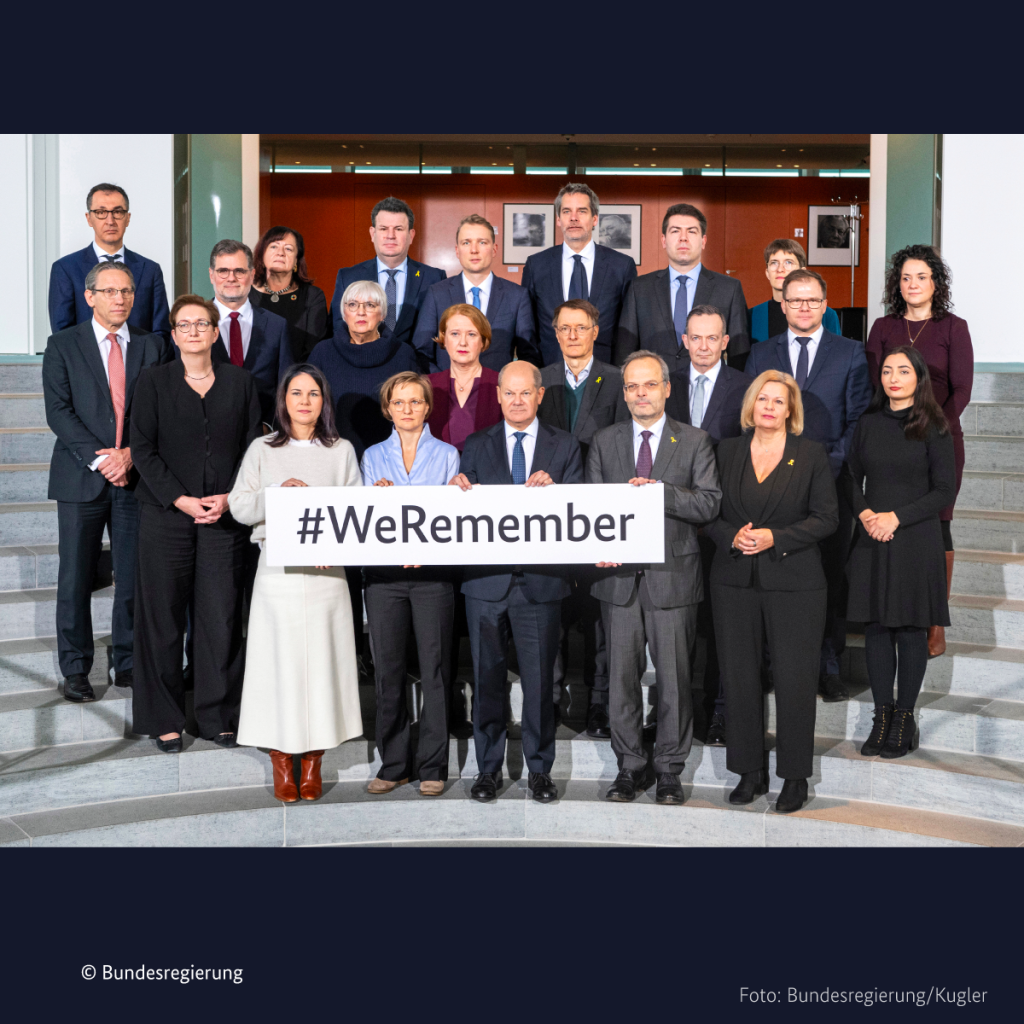 Zu sehen ist ein Gruppenbild des Kabinetts mit Bundeskanzler Olaf Scholz. Der Bundeskanzler, die Bundesaußenministerin Annalena Baerbock und der Beauftragte für Antisemitismus, Felix Klein, sowie Franziska Brantner halten ein Schild, auf dem steht: #WeRemember.