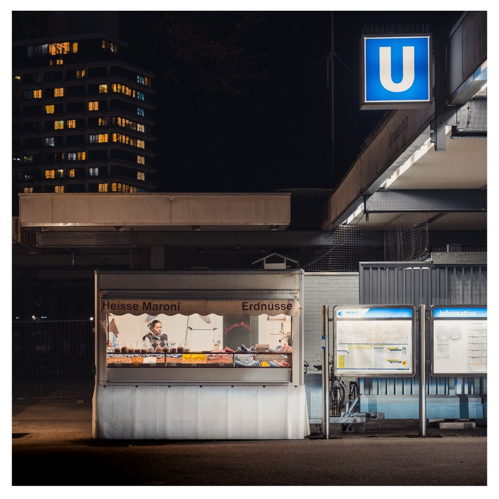 Szene an einer U-Bahnhaltestelle bei Nacht. Im Vordergrund ein Verkaufsstand, hinten ein großes Wohnhaus. 