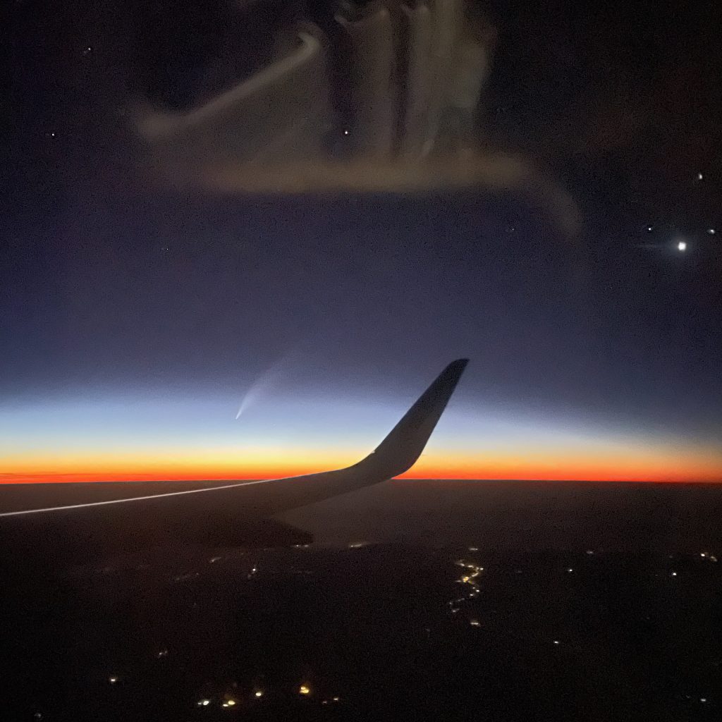 A view from an airplane window during twilight, showing the wing silhouetted against the sky. A comet –a white elongated smudge– hovers above the horizon, surrounded by scattered stars. Below, faint city lights dot the dark landscape.