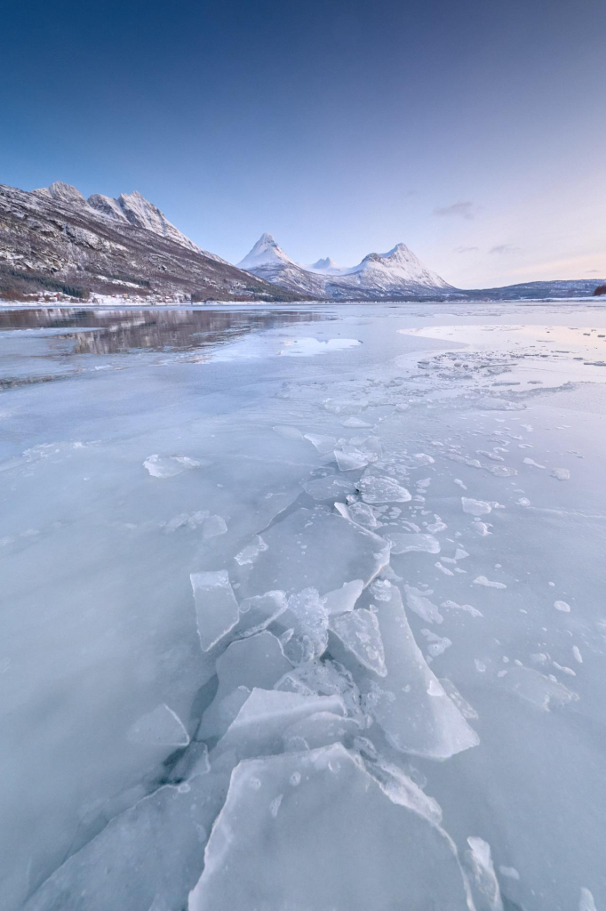 Fjord ice cracking in the foreground, with snow covered mountains in the background.