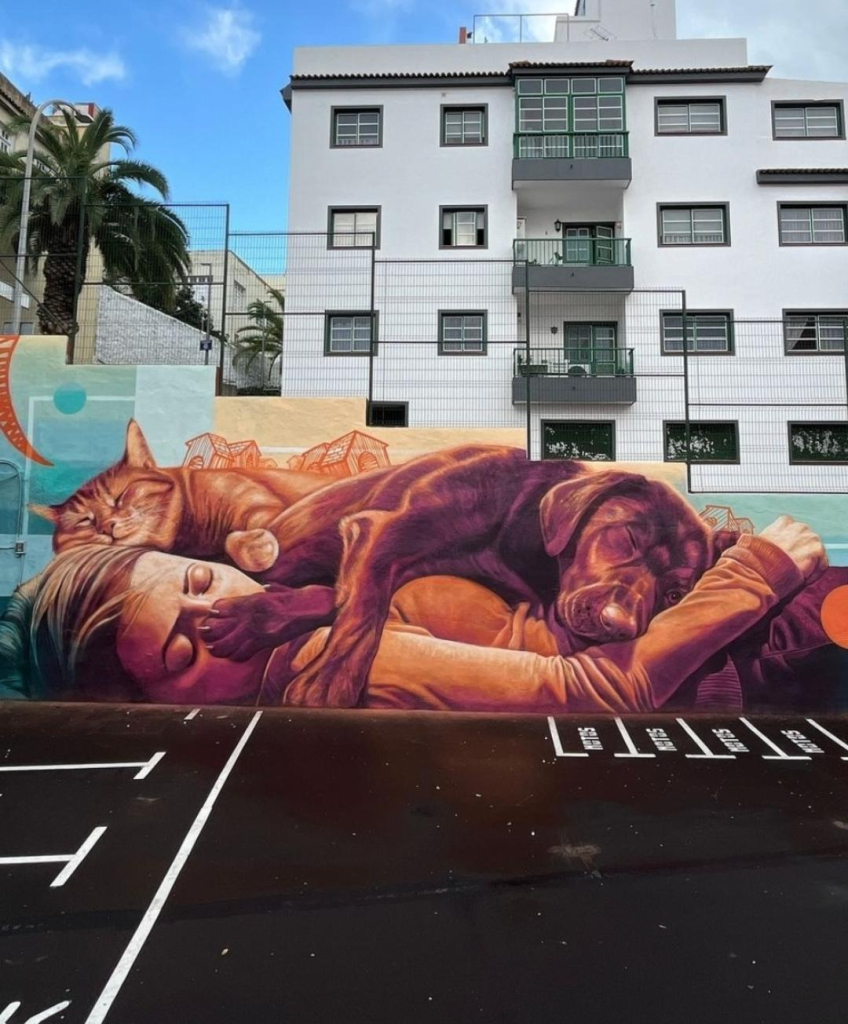 Streetartwall. A very cute mural of a sleeping woman with her pets was sprayed/painted on a long street wall. Painted entirely in shades of orange and brown, you can see a young blonde woman in a t-shirt and jeans lying on the ground sleeping. A sleeping large brown dog lies on top of her, its paw resting directly on the girl's nose. The trio is completed by an orange striped cat lying on and around the young woman's head, sleeping peacefully next to the dog. A row of simply drawn houses above the three symbolizes home and cohesion for all, human and animal. (In the photo you can see a paved parking lot in front and some houses behind it)