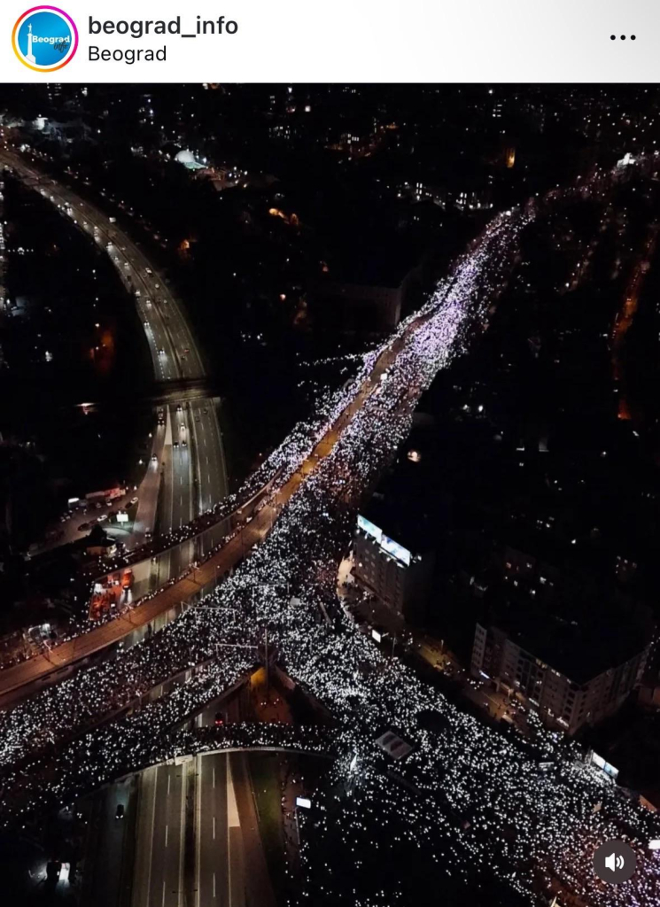 Luftaufnahme einer nächtlichen Protestveranstaltung in Belgrad, Serbien. Zwei große Straßen, die sich an einer Kreuzung treffen, sind von einer riesigen Menge Menschen komplett gefüllt, die mit ihren eingeschalteten Handylampen leuchten. Diese unzähligen kleinen Lichtpunkte erzeugen den Eindruck eines leuchtenden Flusses, der sich durch die Stadt bewegt.

Links im Bild ist eine mehrspurige Schnellstraße zu sehen, die nur spärlich befahren ist, da der Verkehr offenbar wegen der Veranstaltung umgeleitet wurde. Auf der rechten Seite und im Hintergrund sind Gebäude mit leuchtenden Fenstern und Werbetafeln sichtbar. Die Szene wird von den Lichtern der Stadt und der Masse an Protestierenden dominiert, was eine eindrucksvolle und friedliche Atmosphäre schafft.

Das Bild vermittelt die Wirkung einer großen, organisierten Menschenmenge, die ihre Botschaft in einer kraftvollen, symbolischen Weise ausdrückt, indem sie die Dunkelheit mit Licht durchbricht.