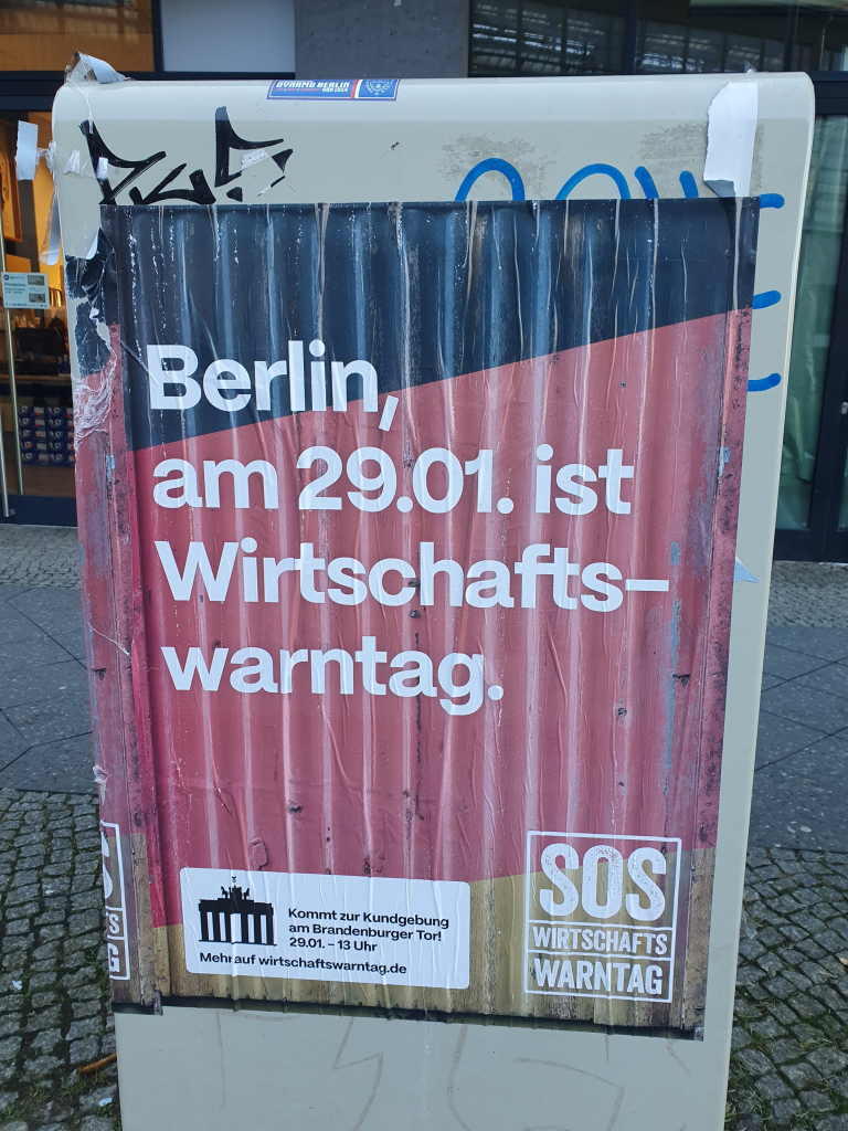 INSM-Plakat an einem Stromkosten, Aufschrift: Berlin, am 29.1. ist Wirtschaftswarntag. Logo der Kampagne SOS Wirtschaft. Im Hintergrund die Farben schwarz-rot-gold.