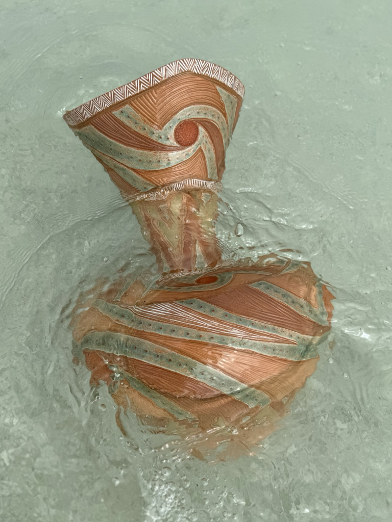 A large ceramic vase with a voluminous, bottom portion, narrow waist and flared neck, at the beach washed over by clear blue ocean water. The vase is intricately decorated with earth tones, and a light aqua glaze patterns swirl around the vase, just as the waves and water do.