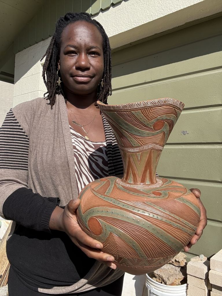 Osa Atoe holding the same vase, standing in front of her pottery studio in the sunshine. The vase is 17” tall and about 12” diameter at its widest point. 