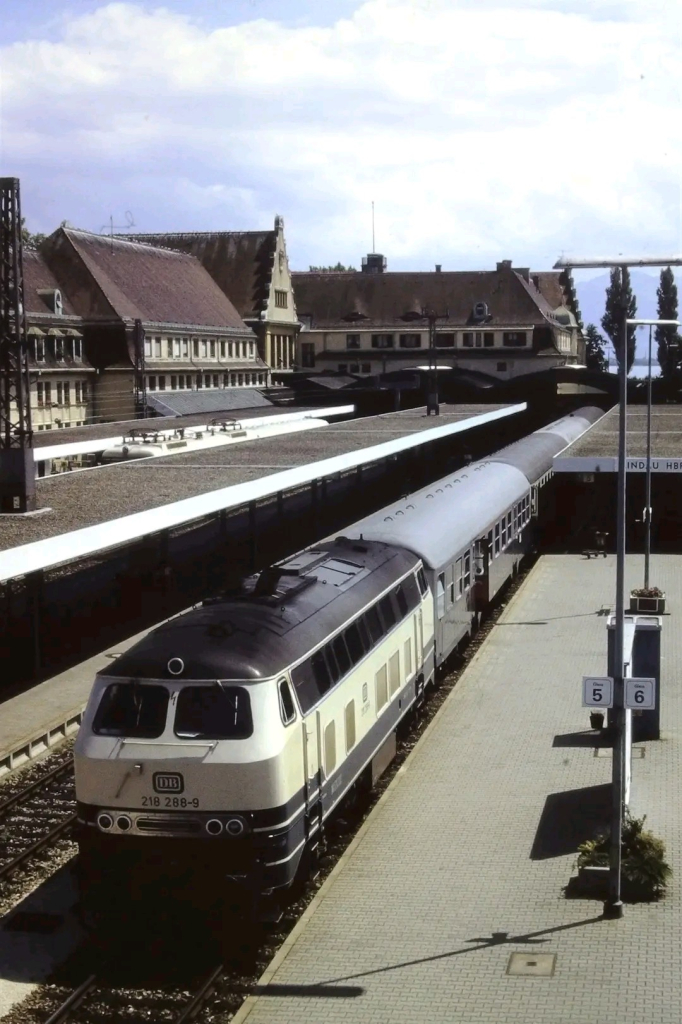 Ein langer Reisezug verlässt einen Bahnhof, gezogen von zwei Diesellokomotiven der Baureihe 218 in beige-blauer Farbgebung. Die Loks fahren in Doppeltraktion und stoßen dunklen Abgasrauch aus. Der erste Wagen des Zuges ist auffällig orange, während die weiteren Wagen eine klassische Schnellzuglackierung in Rot und Beige haben.

Der Bahnhof besteht aus mehreren Gleisen mit Bahnsteigen, und in der linken Bildhälfte stehen abgestellte Reisezugwagen in blauer Farbgebung auf Nebengleisen. Im Hintergrund sind niedrige Häuser mit roten Dächern zu sehen, dahinter erstreckt sich eine hügelige oder bergige Landschaft. Die Szenerie wirkt sommerlich, mit klarem Himmel und grünen Bäumen entlang der Gleise.

