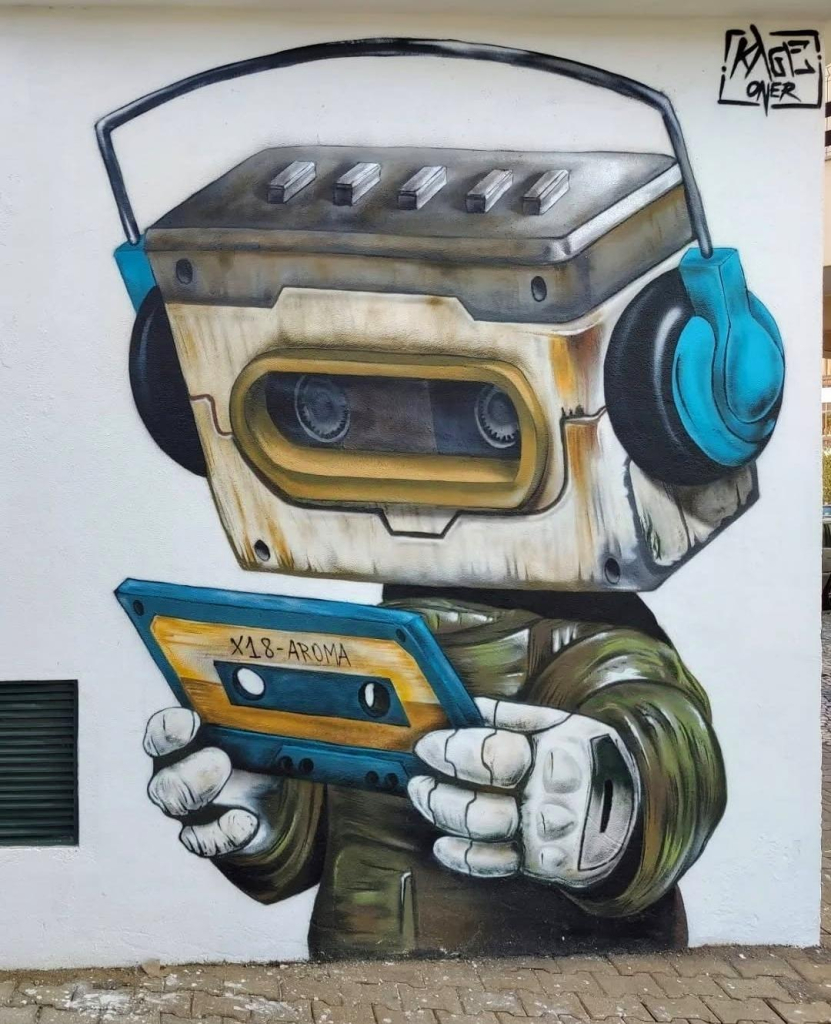 Streetartwall. An original mural with a very musical comic-style figure has been sprayed/painted on a white house wall. The figure is an old Walkman, dressed in an olive-colored jacket and with metal hands. He has green headphones on and is holding an old music cassette in his hands. On the cassette is written X18 Amora (a HipHop/Rap Music Producer). There is already a cassette in the cassette compartment with round holes that look like his eyes. This cute cassette man makes you want to press play immediately.