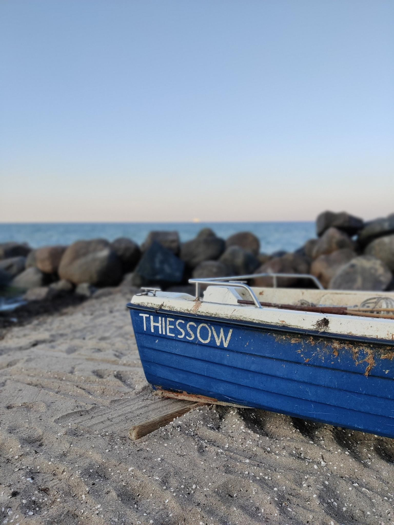 Ein blau/weißes Boot liegt am Strand. Dahinter einige große Felsen als Wellenbrecher vor dem Wasser. Es ist wolkenlos. Im Sand liegen einige Muscheln. Das Boot trägt den Namen "Thiessow"