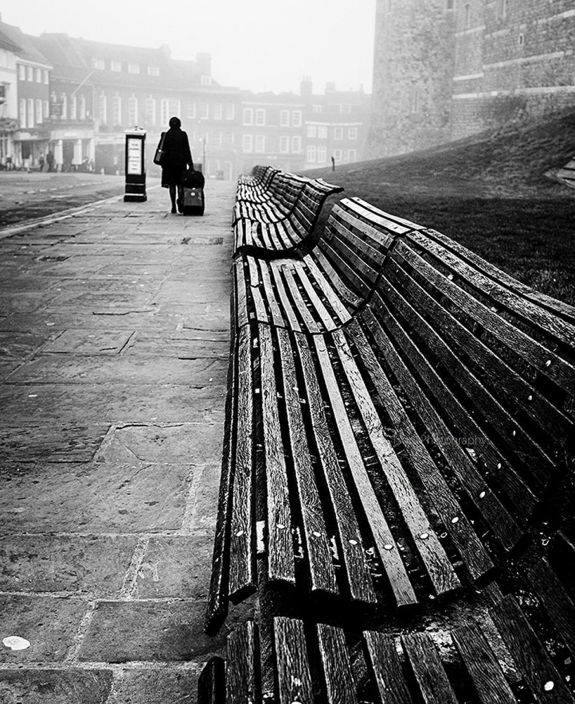 Photography. A black and white photo of a street scene in foggy London. The photograph shows a seemingly endless row of interconnected, curved wooden benches stretching across the entire image on the right. It appears to be early morning. Buildings can be seen in the background, blurred in a misty haze, and you can almost feel the wetness and a certain melancholy.  From behind, a person in a dark winter coat and cap can be seen walking along the stone sidewalk towards the fog, carrying a wheeled suitcase and a handbag. The photo focuses on the benches, which become smaller and smaller and create a diffuse atmosphere that wants to continue telling the story of the person on her way.