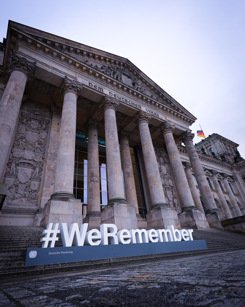 Auf einem Foto ist die Vorderseite des Reichstagsgebäudes zu sehen, davor eine Plastik bestehend aus großen weißen leuchtenden Lettern: „#WeRemember“.