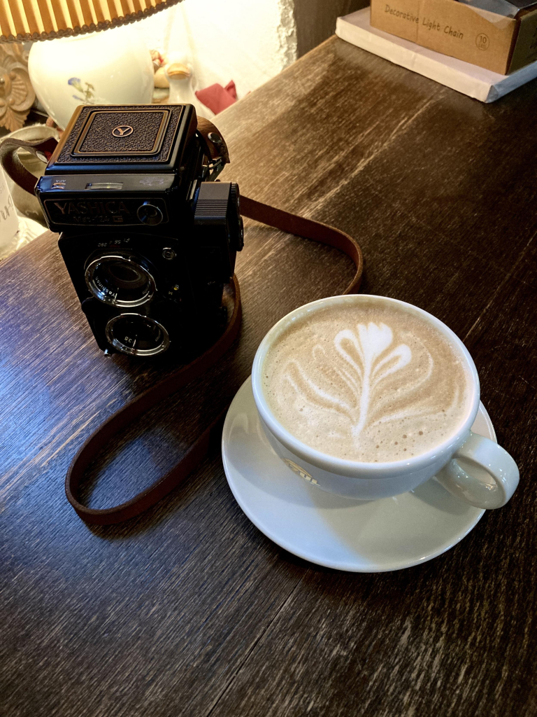 Eine alte analoge Kamera, eine sogenannte TLR mit 2 Linsen steht auch einem dunkelbraunen Tisch, daneben ein Capucchino. Im Hintergrund ist noch eine alte Lampe zu sehen.

