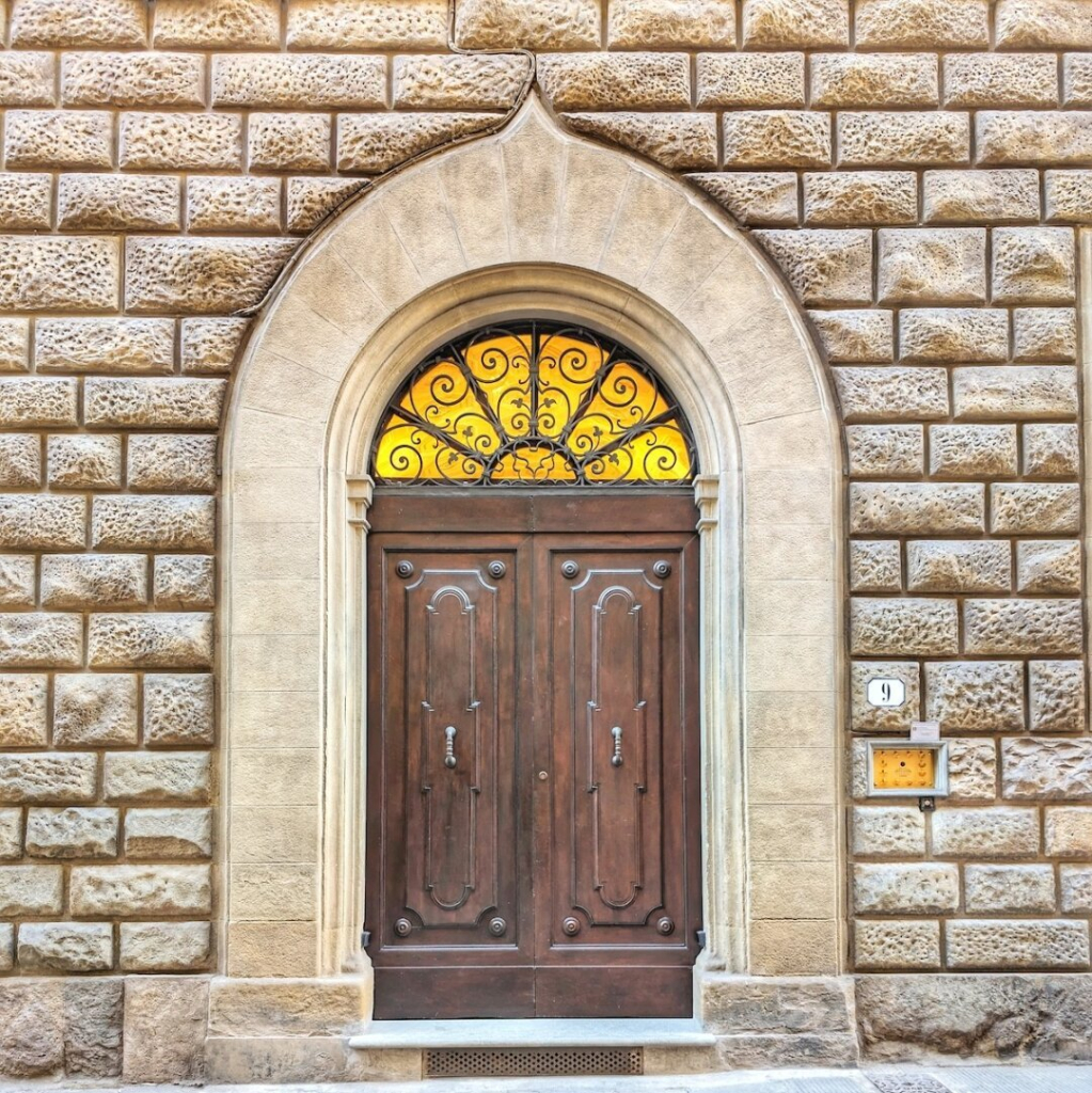 A grand wooden door, rich brown and accented with lighter decorative panels, stands within a wall of large, light sandy bricks. Above the door, a fanlight of ornate wrought iron glows with a warm yellow light. This arched window is framed by the same pale stone that surrounds the door, contrasting with the rough texture of the brickwork. A small rectangular sign to the right of the door bears the number "9".