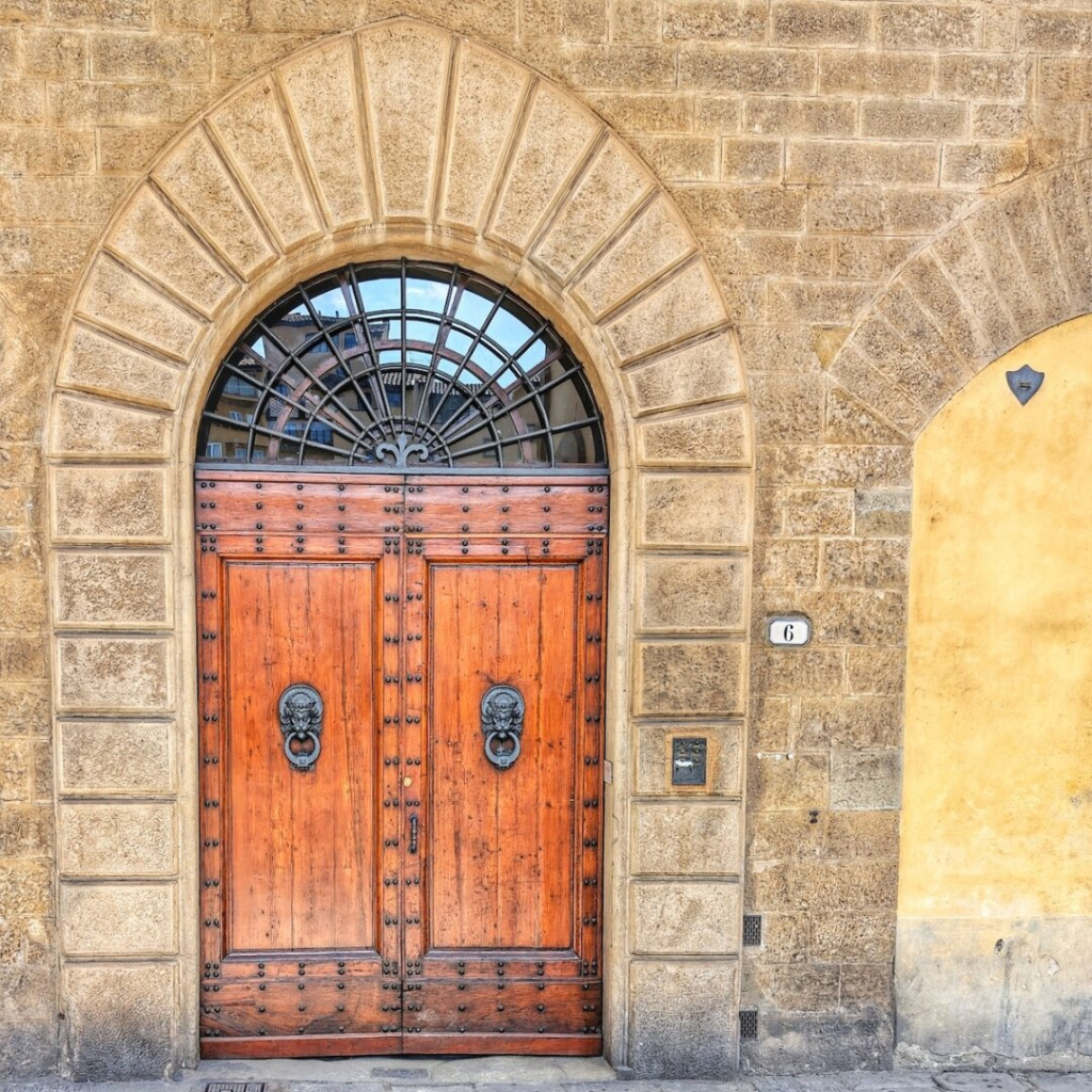 A close-up photograph captures a brown double door nestled in an aged, light brown brick wall. The door, crafted from wood, is adorned with numerous dark metal studs and features two lion head door knockers. Above the door, a semi-circular window is framed by a matching archway. The window is divided into small panes by a grid of metal bars, with a central design reminiscent of a flower or a starburst. To the right of the door, a small black sign displays the white numeral "6". The brick wall extends beyond the door, showing variations in color, with the section to the right of the door appearing to be a pale yellow. The bricks themselves are uneven in shape and size, adding to the building's rustic charm. The photograph's lighting suggests an overcast day or a shaded location, as the colors are muted and there are no harsh shadows.