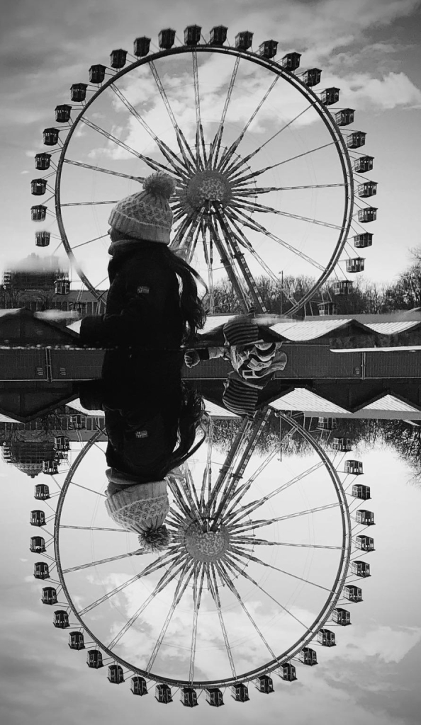 A black and white image of a ferris wheel with a reflection in water. In the foreground, a person wearing a knitted hat and coat walks alongside the ferris wheel, creating a striking visual contrast with the reflection below.