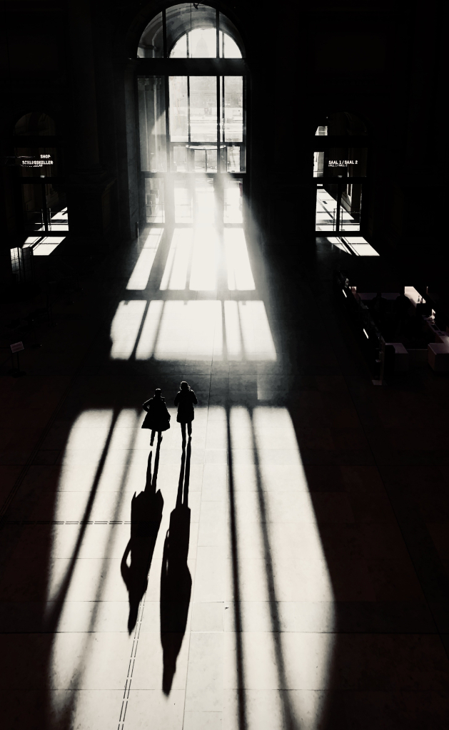 Silhouettierte Figuren stehen in einer sonnenbeschienenen Halle mit langen Schatten auf dem Boden, große Fenster im Hintergrund.