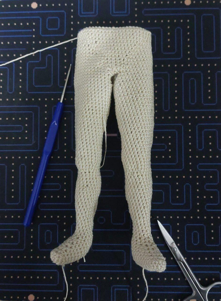 An image of semi-realistic crocheted legs and part of the torso for an amigurumi doll. They're made out of thin beige yarn. The image also features a small crochet hook and scissors.