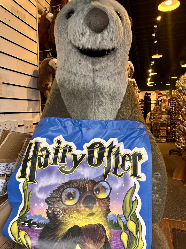 A photo of a large otter mascot holding a shirt that says Hairy Otter. The otter character is wearing ‘the boy wizard’ style glasses 