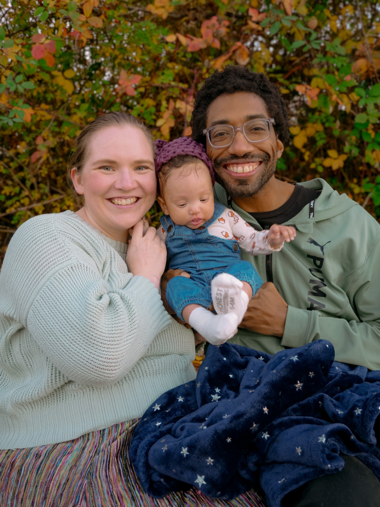 Two smiling adults are sitting outdoors and holding a 3-month-old baby between them. On the left is a white nonbinary adult, on the right is a Black man with glasses. The baby is light-skinned, wearing denim overalls, and is looking down with one hand up.