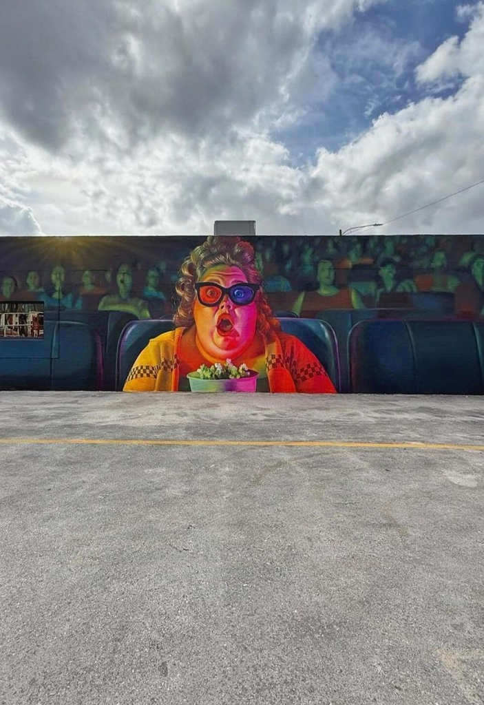 Streetartwall. A mural with a movie scene was painted on a long exterior wall. The colorful picture shows a slightly corpulent lady in an orange vest sitting in the front row of a movie theater. She has a curly hairstyle, wears 3D glasses, holds a box of popcorn in her hand and has her mouth wide open. Something wild must be happening on the screen. Behind her, countless moviegoers are sitting in their seats, feeling the same way. But the focus is on Madame, because the background is only blurred. Only part of the very long wall and the parking lot in front of it can be seen in the photo.