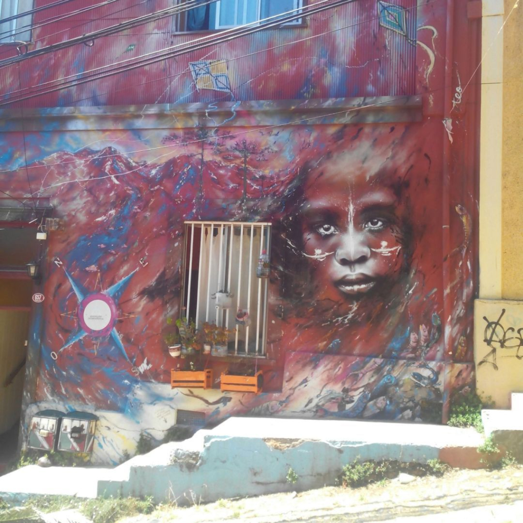 Photo of the front of a house in Valparaíso, Chile. The wall is painted in Red colors with some touch of blue. There is a moutain painted in the style of a galaxy. The face of a black person is painted.