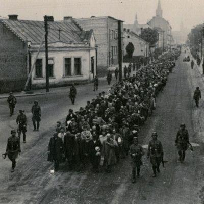 A historical black and white photo depicting a large group of people being marched down a city street, with some buildings visible on either side.