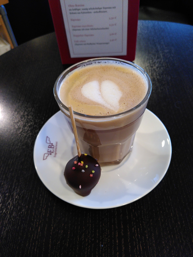 Auf einer weißen Untertasse mit dem Logo des Cafés auf der linken Seite (die Großbuchstaben *E und B*, eingefasst von zwei Flügeln; darunter steht *Engelsbäckerei*) steht ein halbhohes Glas mit einer Mischung aus Espresso und heißer Vollmilchschokolade namens Schokopresso. Die Oberfläche ist mit einem Herz aus Milchschaum dekoriert. Davor thront ein runder Kuchen am Stiel. Dieser ist in Edelbitterschokolade getaucht und ist mit einigen bunten, kleinen Zuckersternen  dekoriert. 
Im Hintergrund ist ein Teil der in dunkelrot gefassten Getränkekarte zu sehen.