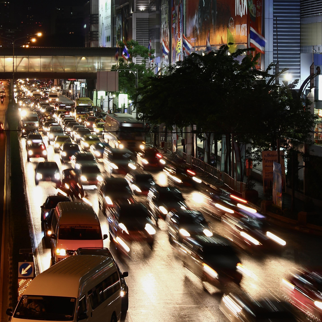 das quadratische bild zeigt eine straßenszene mit vielen autos in der dunkelheit.
von einer brücke fotografiert ist eine mehrspurige straße zu sehen. autos kommen einem entgegen. da es eine langzeitbelichtung ist, sind die autos, die vorne in bewegung sind, in unschärfe zu sehen, während in der straßenflucht nach hinten die fahrzeuge scharf abgebildet werden. rechts neben der straße ist ein hohes gebäude mit fassadenwerbung. davor sind einige bäume an der straße. weiter hinten ist ein überdachter fußgänger*innenüberweg zu sehen.
die aufnahme entstand in bangkok.