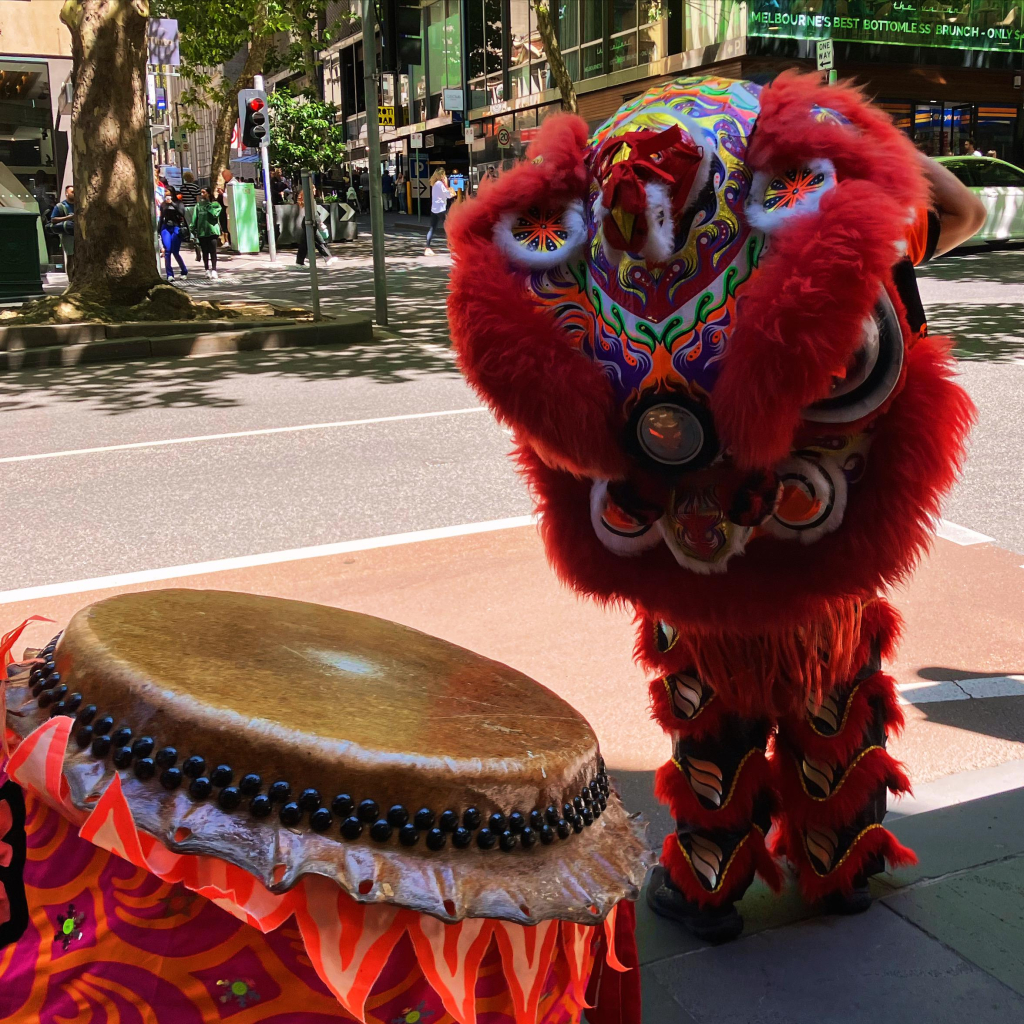 Someone in a mostly red lion dance costume stands next to a Chinese drum at a bus stop 