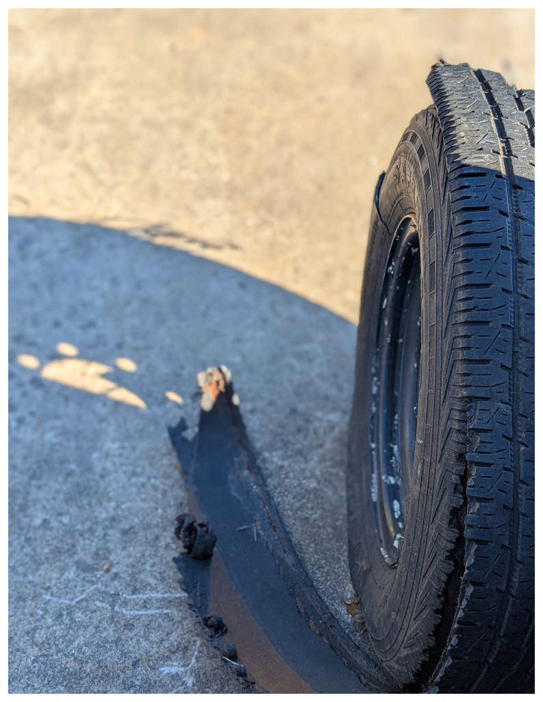 high-angle view of a vehicle tire with recapped material that has torn and is falling away. the background is a shadow of the tire on concrete.