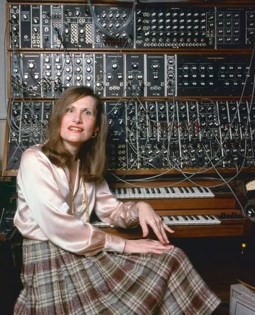 Wendy Carlos, musician, sitting before a huge devise with piano keys and an enormous amount of plugs and cables. She wears a brownish skirt and pink/light brown blouse. 